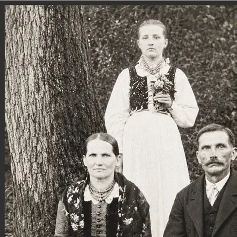 Rodzinny portret: kobieta z kwiatami w haftowanym gorsecie i dwoje starszych ludzi w strojach regionalnych.